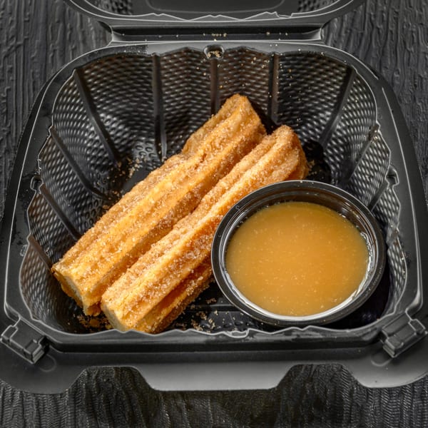 Two Sabor Tacos churros, coated in cinnamon sugar, with a side of caramel dipping sauce in a black container.