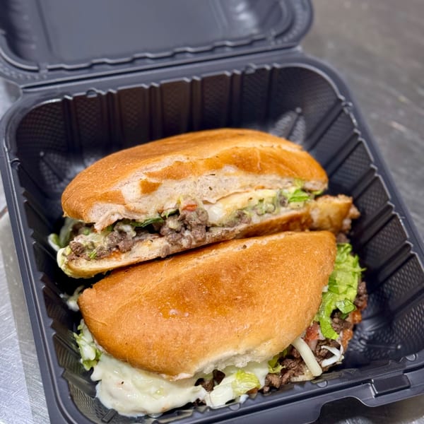 Two Sabor Tacos tortas with ground beef, lettuce, and white sauce in a black to-go container.