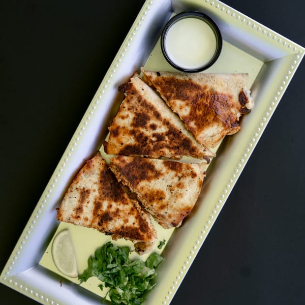 Four crispy, golden-brown quesadilla wedges with a side of crema, lime, and cilantro on a tray.